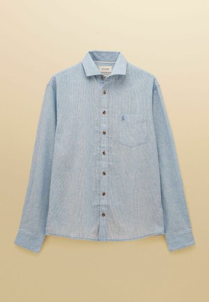 Svetlomodrá košeľa s dlhým rukávom na gombíky s hnedými gombíkmi a náprsným vreckom s malým vyšitou logom na béžovom pozadí.