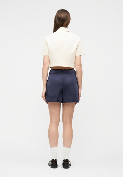 Γυναίκα που στέκεται με την πλάτη στραμμένη, φορώντας ένα κρεμ κοντομάνικο cropped τοπ, σκούρο μπλε σορτς ψηλόμεσο, λευκές κάλτσες και μαύρα μοκασίνια σε λευκό φόντο.