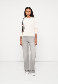 Kremowy sweter w warkocze, szare dżinsy z szerokimi nogawkami, białe trampki i mała torebka w wzory. Prosty, codzienny strój o luźnym kroju.