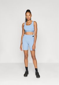 Svijetloplavi atletski crop top s širokim naramenicama i logom na elastičnoj traci, u kombinaciji s visokim hlačicama koje odgovaraju. Ima glatku teksturu i tijesno pristajanje.