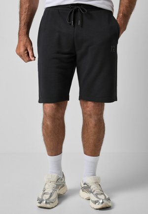 Uomo con pantaloncini neri con coulisse, calzini bianchi alti e scarpe da ginnastica bianche e grigie, in piedi su uno sfondo chiaro e uniforme.