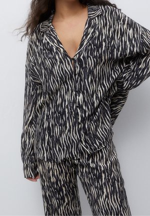 Femme portant une chemise à boutons noire et blanche à rayures zébrées avec un pantalon ample assorti, debout avec une main sur la hanche.