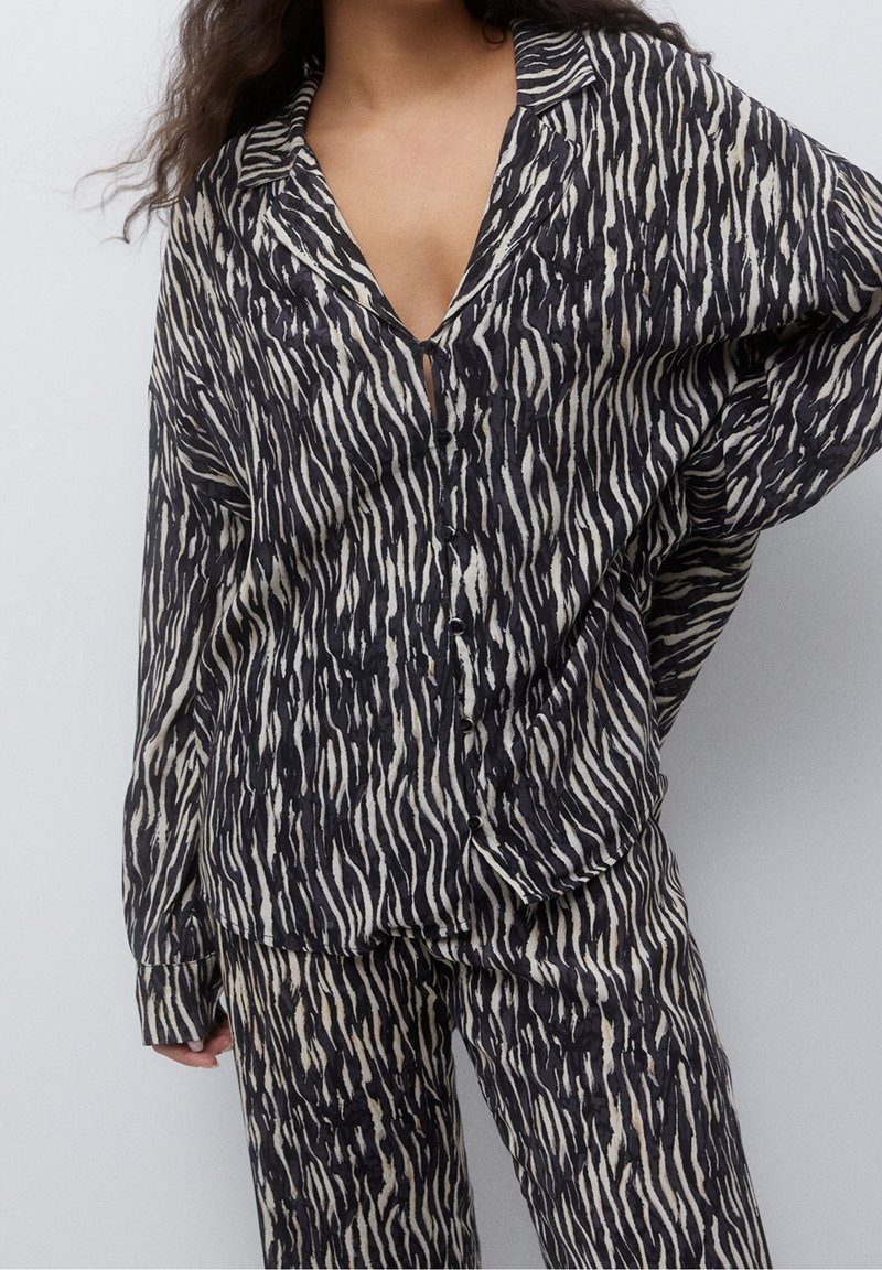 Femme portant une chemise à boutons noire et blanche à rayures zébrées avec un pantalon ample assorti, debout avec une main sur la hanche.