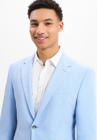 Lichtblauwe op maat gemaakte blazer met een gladde textuur, een enkele knoopsluiting en notch-revers. Draagt op een witte overhemd met kraag.