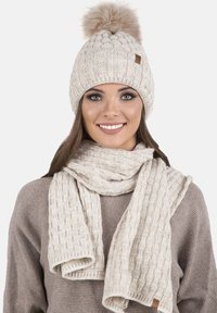 Gorro de punto beige con un pompón y bufanda texturizada a juego. Cuenta con un patrón acanalado y una pequeña etiqueta de cuero en el gorro.