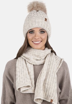 Béret en tricot beige avec un pompon et une écharpe texturée assortie. Présente un motif côtelé et une petite étiquette en cuir sur le chapeau.