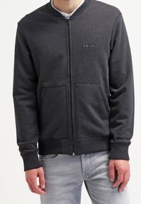 Homme portant un sweat-shirt noir zippé avec le logo « Bench. » sur la poitrine et un jean gris clair, debout les mains détendues le long du corps.