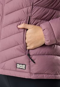 Hand in der Reißverschlusstasche einer gesteppte mauvefarbenen Jacke mit Denim Factory Etikett, die gepolsterte Stofftextur und Nahtdetails zeigt.
