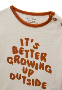 T-shirt en coton beige clair avec un col rond bordé d'orange. Texte en lettres brunes grasses : "IL EST MEILLEUR DE GRANDIR DEHORS."