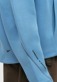 Ανοιχτό μπλε φούτερ με μακριά μανίκια, με μαύρο τυπωμένο λογότυπο στο μανίκι και ραφές σε αντίθετο χρώμα. Μαλακή υφή υφάσματος.