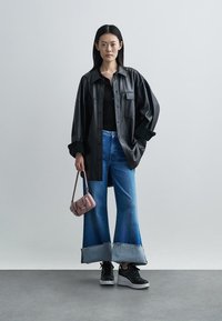 Schwarzes, übergroßes Hemd aus Leder, schwarzes Oberteil, weite blaue Flared-Jeans mit ausgefransten Säumen, schwarze Sneakers und eine kleine pinke Handtasche.