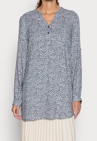 Blouse grise à manches longues avec un décolleté en V, présentant un motif specklé multicolore et des boutons à l'avant. Tissu léger.