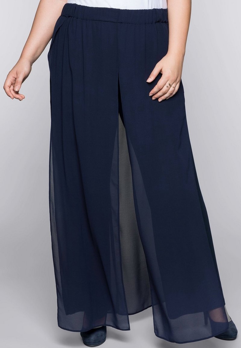 Femme portant un pantalon large en tissu transparent bleu marine et des chaussures foncées, debout, les mains détendues le long du corps sur un fond gris clair.