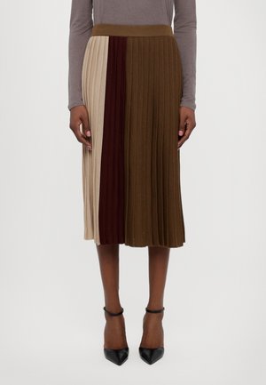 Kobieta nosząca plisowaną spódnicę midi z pionowymi paskami w kolorach beżowym, ciemnobrązowym i oliwkowozielonym, w połączeniu z czarnymi szpilkami na kostkę.