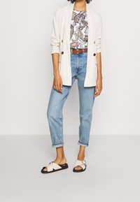 Blazer beige claro sobre una blusa blanca con estampado, jeans de mezclilla azules y sandalias rosas con suela negra y detalles de hardware plateado.