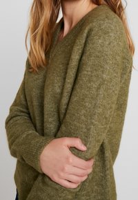 Mujer con cabello largo y rubio, vistiendo un suéter de punto verde oliva, sosteniendo su brazo izquierdo con la mano derecha.