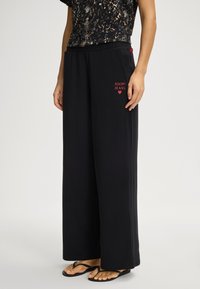 Svarta byxor med vida ben tillverkade av mjukt tyg, med elastisk midjeresår och röd "JOOP! JEANS"-logga framtill för varumärkesprofilering.