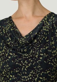 Czarna tkanina z zielonymi i żółtymi wzorami przypominającymi liście; charakteryzuje się drapowanym dekoltem i miękką teksturą. Zbliżenie na projekt odzieży.