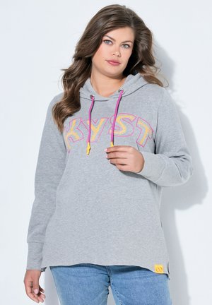 Kvinde med langt brunt hår iført en grå hættetrøje med lyserøde snore og teksten "KYST", kombineret med blå jeans, foran en hvid baggrund.