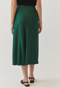 Gonna verde di media lunghezza realizzata in tessuto liscio, con un design fluido e una vita elastica. Indossata con sandali neri piatti.