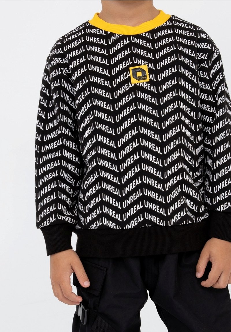 Sweatshirt noir avec le texte « UNREAL » en blanc, doté d'un col rond jaune et d'un petit logo. Le texte est disposé en motif en chevron.