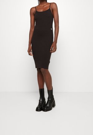 Woman wearing a fitted black knee-length dress with thin straps and black heeled ankle boots, standing against a plain white background.