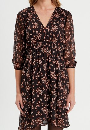 Robe noire avec motif floral rose, décolleté en V, manches trois-quarts transparentes et taille froncée, portée par une personne debout.