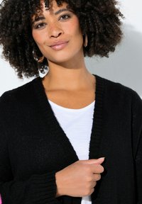 Sort strikket cardigan med V-hals og ribbede ærmekanter, båret over en hvid skjorte. Tyk, tekstureret stof med en afslappet pasform.