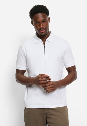 Witte poloshirt met een ritssluiting bij de kraag, korte mouwen en een gladde textuur, gecombineerd met kaki broek. Model staat met de handen gevouwen.