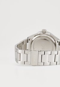 Ασημένιο ρολόι από ανοξείδωτο ατσάλι με βραχιόλι από λινκ, γυαλιστερό φινίρισμα, στρογγυλή θήκη και ορατό εγ engraved λογότυπο στη γλώσσα του κλιπ.