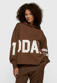 Sweat à capuche marron surdimensionné avec lettrage blanc en gras "10DA" et accents à rayures sur les manches, fabriqué en tissu doux et texturé.