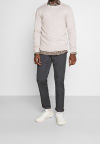 Pull en tricot beige clair porté sur une chemise à manches longues à imprimé floral foncé, associé à un pantalon gris foncé et des baskets blanches. Tenue décontractée moderne.