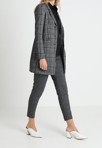 Blazer en écossais gris à coupe ajustée, associé à un pantalon gris fuselé et des mules blanches pointues, vu de profil.