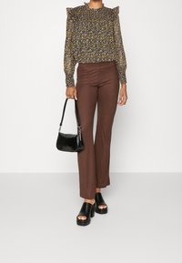 Blusa a stampa floreale con scollatura arricciata, pantaloni marroni a coste a vita alta e sandali neri con tacco. Il modello tiene una piccola borsa nera.