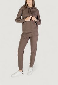 Mujer vestida con una sudadera marrón de la marca EA7 y pantalones deportivos a juego, de pie con las manos ajustando los cordones de la sudadera, llevando zapatillas blancas.