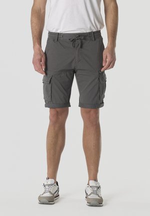 Hombre vistiendo pantalones cortos cargo grises con dobladillos enrollados, zapatillas blancas y una camiseta blanca, de pie frente a un fondo liso.