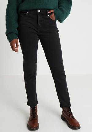Persoon met zwarte high-waisted jeans, donkergroene trui met lange mouwen en bruine leren veterlaarzen, staand op een effen achtergrond.