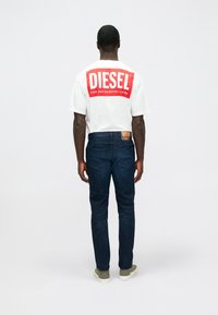 Λευκό βαμβακερό t-shirt με μεγάλο κόκκινο λογότυπο "DIESEL" στην πλάτη. Συνδυασμένο με σκούρο μπλε τζιν παντελόνι και γκρι αθλητικά παπούτσια.