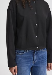 Black cropped jacket made from smooth fabric, featuring a button closure with gray buttons and long sleeves, paired with blue denim jeans.