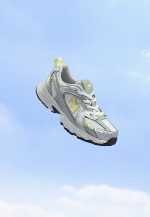 Γκρι και λευκό παπούτσι τρεξίματος New Balance με κίτρινες πινελιές που αιωρείται μπροστά σε καθαρό μπλε ουρανό.