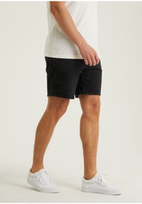 Mustat farkkushortsit, joissa on rento istuvuus, polven ylle ulottuva pituus ja klassinen viisi-tasku design, yhdistettynä valkoisiin lenkkareihin ja kevyeseen paitaan.