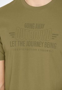 Oliivinvihreä puuvillapaita, jossa on pyöreä kaula-aukko. Siinä on musta graafinen teksti: "MENOSSA MATKALLE, ANNA MATKAN ALKAA, KOHDE TUNNETON."