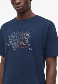 Homme portant un t-shirt bleu marine avec un graphisme représentant plusieurs silhouettes courant, contourées en blanc et orange avec le texte "new balance".