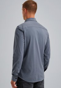 Hombre con camisa de manga larga azul grisácea visto desde atrás, con un fondo liso gris claro.