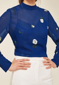 Himmelbluse aus transparenter Stoff mit hohem Hals, langen Ärmeln und weißen Blumenmustern. Kombiniert mit weißen Hosen, die filigrane Stoffdetails zeigen.