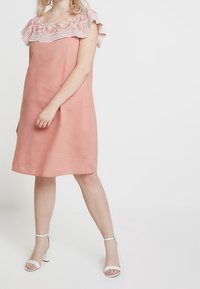 Vaaleanpunainen pellavapuku, jossa on röyhelöinen pitsikaulus, väljä istuvuus ja polvipituinen design. Yhdistetty valkoisiin korkokenkiin.