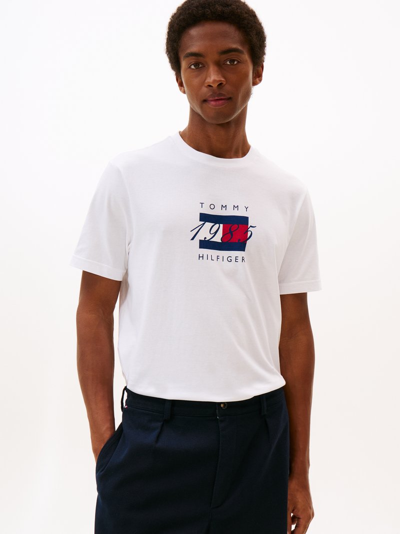 Bílý bavlněný tričko s nápisem "TOMMY HILFIGER" a "1985" v námořnické a červené grafice. Kulatý výstřih a krátké rukávy, kombinované s tmavými kalhotami.