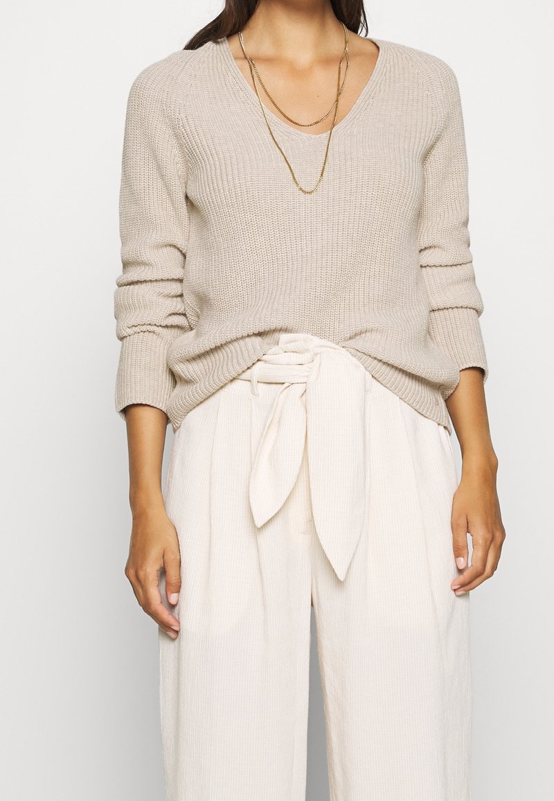 Femme portant un pull en maille beige à col en V, un long collier en chaîne dorée et un pantalon crème taille haute avec une ceinture en tissu nouée.