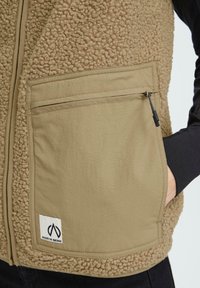 Beige Fleece-Weste mit einer strukturierten, flauschigen Außenseite und einer glatten Stofffront. Verfügt über eine Reißverschlusstasche und ein sichtbares Markenlabel.