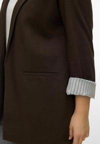 Blazer marron à coupe droite, avec une poche unique et des revers rayés gris retroussés. Texture lisse, design minimaliste.